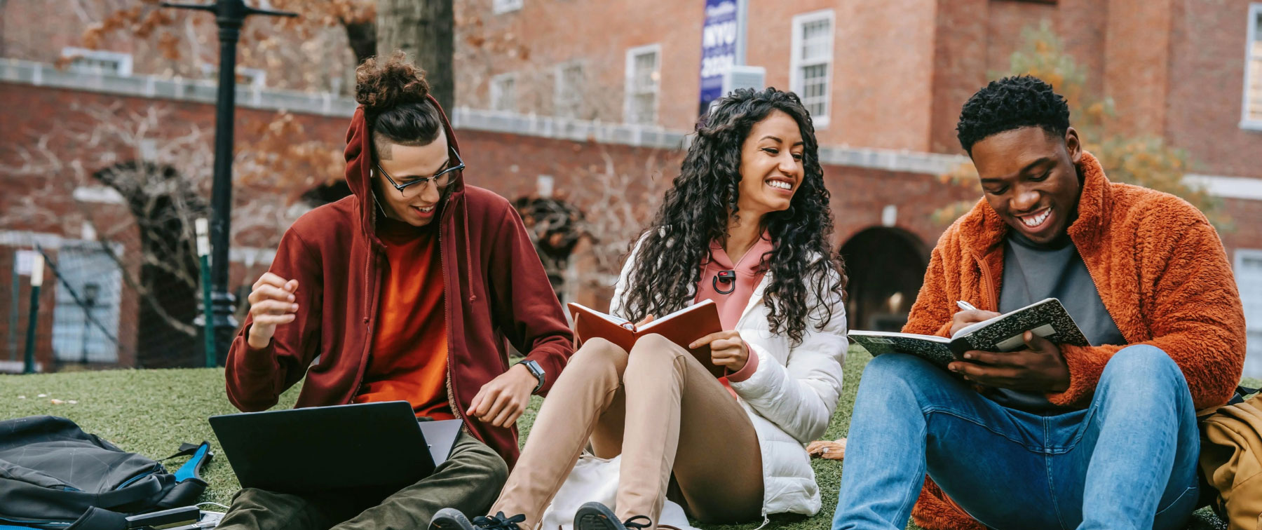 Group of three young college-aged people smiling while reading together outside